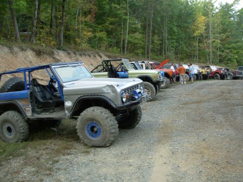1983 Ford Bronco - Multiple modified Ford Broncos are lined up on a dirt and gravel lot near a wooded area.