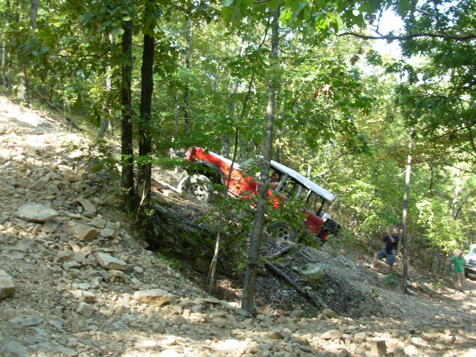 Red white-roofed 4x4 climbing a steep rocky wooded hillside trail, front end raised