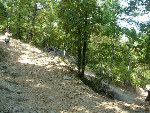 1983 Ford Bronco - An off-road vehicle struggles on a steep, rocky trail surrounded by trees, with another vehicle visible in the distance.