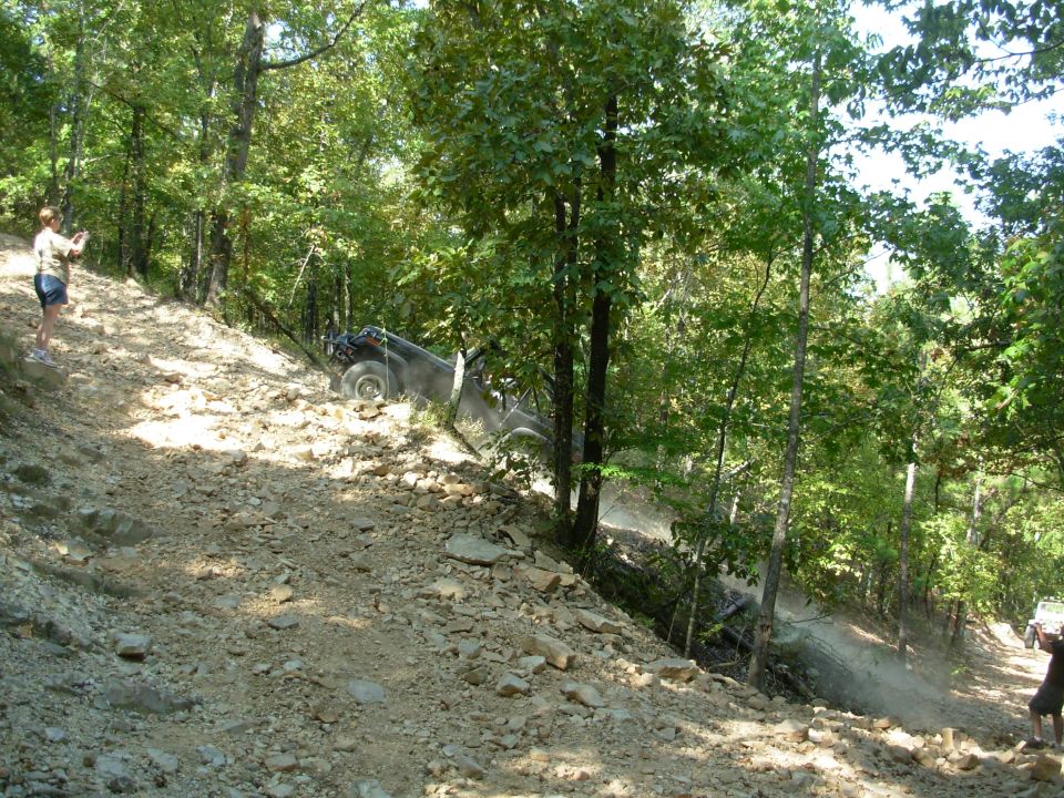 Dark lifted 4x4 climbing a steep rocky wooded off-road trail as spectators watch