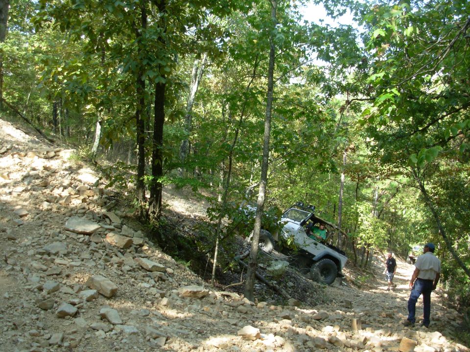 White open-top 4x4 with light bar crawling down a steep rocky wooded trail as spectators watch