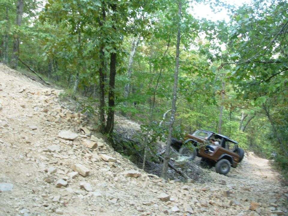Brown topless full-size Bronco with roll bar climbing a loose rock trail in the woods