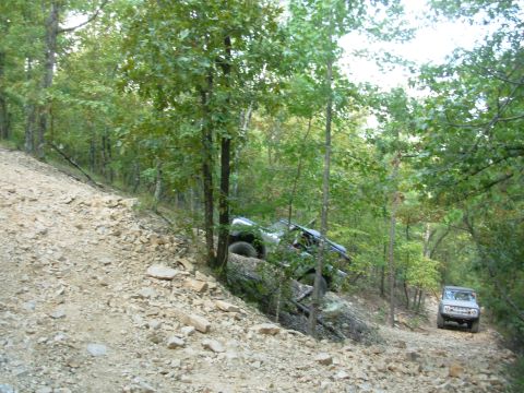 1983 Ford Bronco - Two off-road vehicles navigate a steep, rocky trail through a wooded area, with one vehicle tilted precariously.