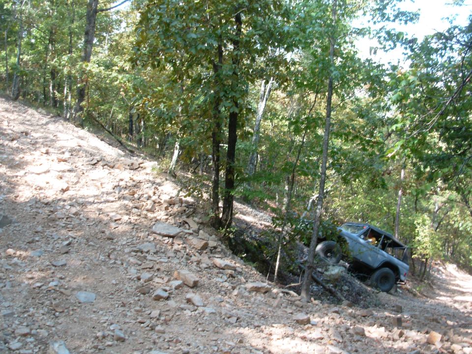 Grey topless Ford Bronco with roll cage climbing a rocky wooded off-road trail