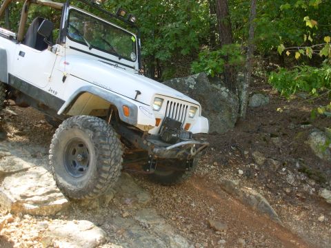 1983 Ford Bronco - White Jeep Wrangler navigates a challenging rocky trail with its front-right tire high in the air.