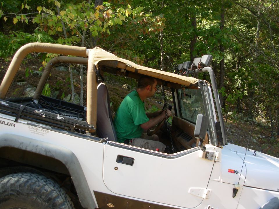 Man driving a white Jeep Wrangler with top folded onto tan roll bars on a trail