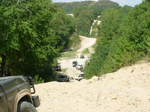 1983 Ford Bronco - A group of SUVs navigates a challenging off-road trail surrounded by lush green trees and hills.