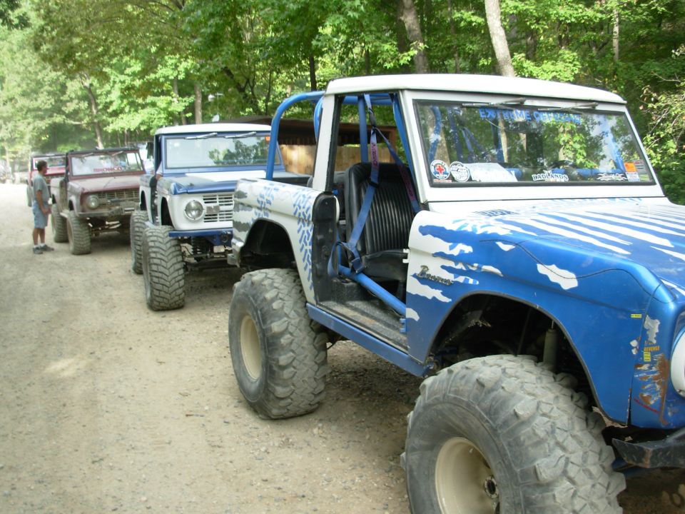 Row of early Ford Broncos on a gravel trail, nearest one blue and white on mud tires