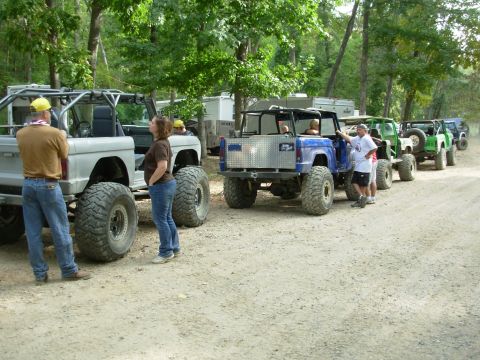 1983 Ford Bronco - A group of off-road vehicles are parked in a line on a dirt path surrounded by trees.
