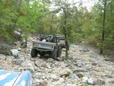 1983 Ford Bronco - Two modified off-road vehicles navigate a challenging, rocky path in a wooded area.