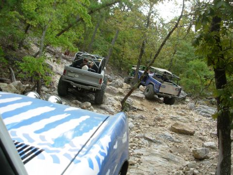 1983 Ford Bronco - Multiple off-road vehicles, including a 1983 Ford Bronco, navigate a challenging rocky terrain on a wooded trail.