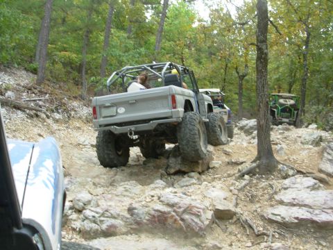 1983 Ford Bronco - Several modified off-road vehicles tackle a challenging rocky trail in a wooded area.