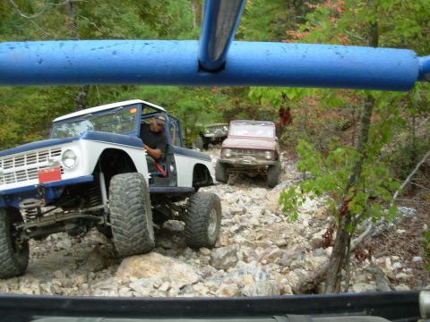 1983 Ford Bronco - Several Broncos navigate a challenging, rocky off-road path through a wooded area.