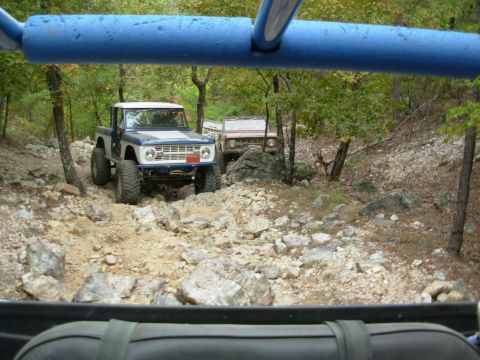 1983 Ford Bronco - Two off-road vehicles navigate a challenging rocky terrain with trees in the background.