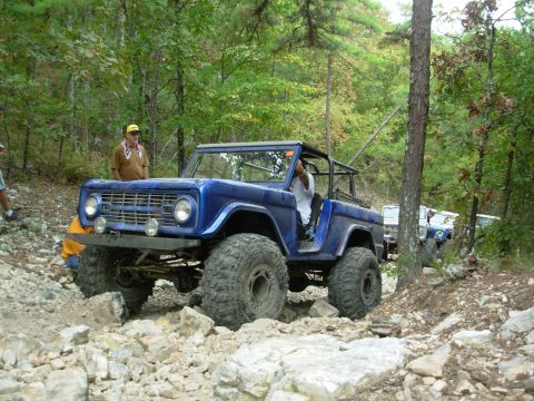 1983 Ford Bronco - A blue SUV with a roll cage is shown on a challenging off-road trail with large rocks.