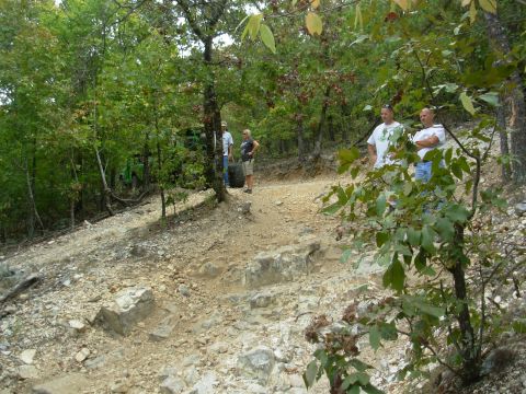 1983 Ford Bronco - Several people stand near a green off-road vehicle on a challenging rocky trail surrounded by trees.