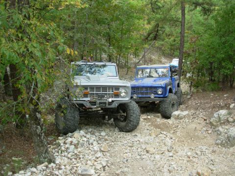 1983 Ford Bronco - Two classic Ford Broncos navigate a challenging, rocky off-road path surrounded by trees.