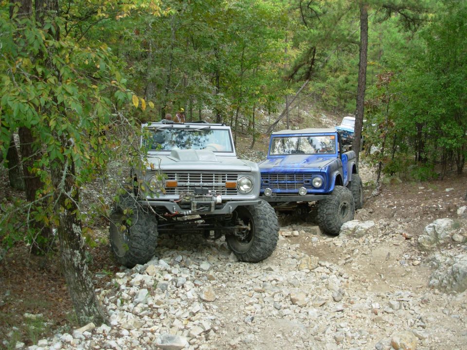 Silver and blue early Ford Broncos, tops off and lifted, parked on a rocky wooded trail