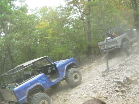 1983 Ford Bronco - Two modified Ford Broncos navigate a steep, rocky trail surrounded by trees.