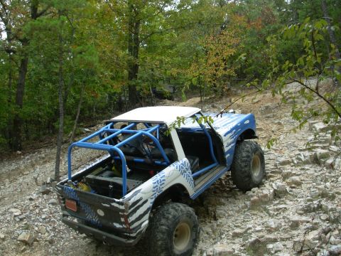 1983 Ford Bronco - This heavily modified SUV navigates a challenging, rocky off-road path surrounded by trees.