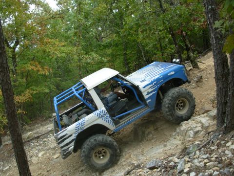 1983 Ford Bronco - Modified off-road vehicle navigates a steep, rocky trail surrounded by trees.