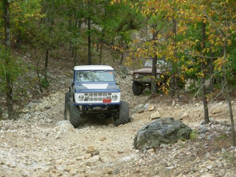 1983 Ford Bronco - Two Ford Broncos navigate a challenging rocky trail through a wooded area.