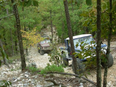 1983 Ford Bronco - Two off-road vehicles traverse a challenging rocky path through a dense forest.