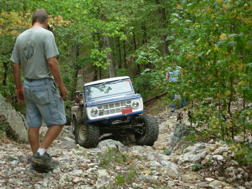 Blue and white early Bronco crawling a rocky wooded trail with a spotter walking ahead