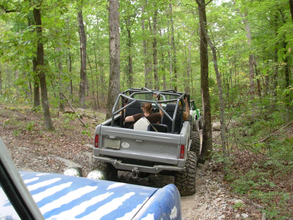 Rear of a silver topless Ford Bronco with roll cage and mud tires on a wooded trail