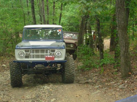 1983 Ford Bronco - Two modified Ford Broncos navigate a wooded trail during an off-road excursion.