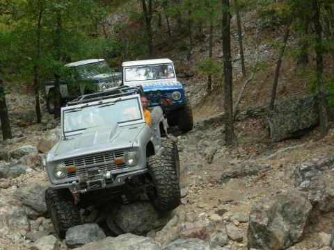 1983 Ford Bronco - Multiple Ford Broncos tackle a challenging, rocky off-road course in a wooded area.