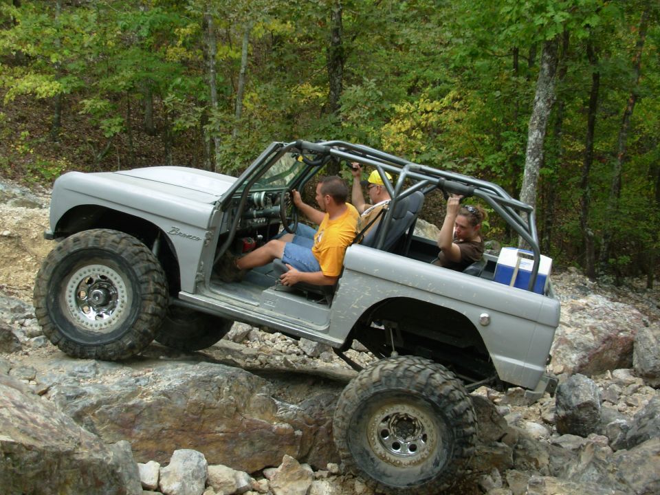 Silver open-top first-gen Ford Bronco with roll cage crawling boulders, three aboard
