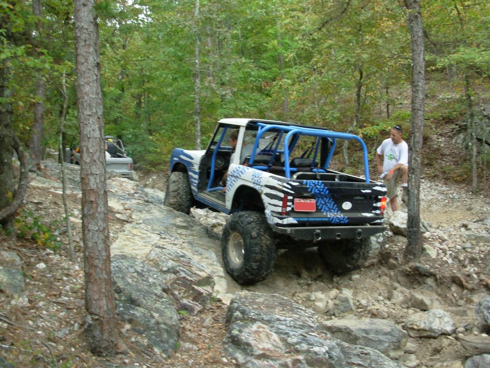 White and blue topless early Ford Bronco with roll cage crawling rocks on a wooded trail