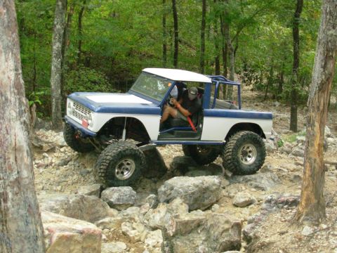 1983 Ford Bronco - A heavily modified Ford Bronco is shown off-roading over rocks in a wooded area.