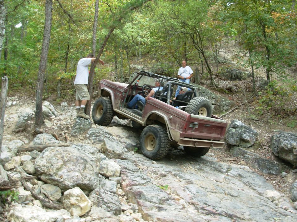 Maroon caged open-top early Ford Bronco crawling down rocks on a wooded trail