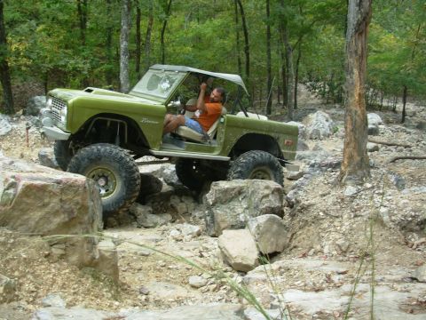 1983 Ford Bronco - A man drives a lifted green Ford Bronco over large rocks on a wooded trail.