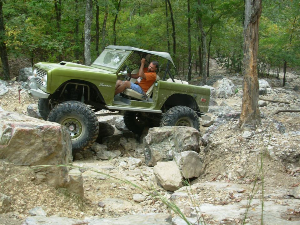 Green doorless early Ford Bronco with white top rock crawling on wooded trail