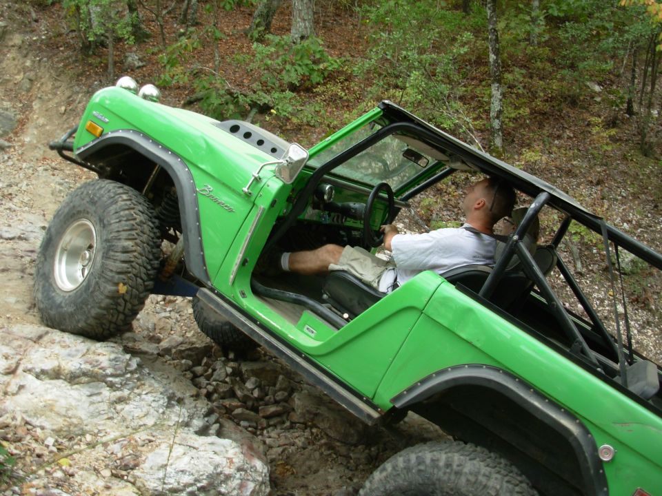 Lime green caged early Ford Bronco tilted on a rock ledge with driver aboard