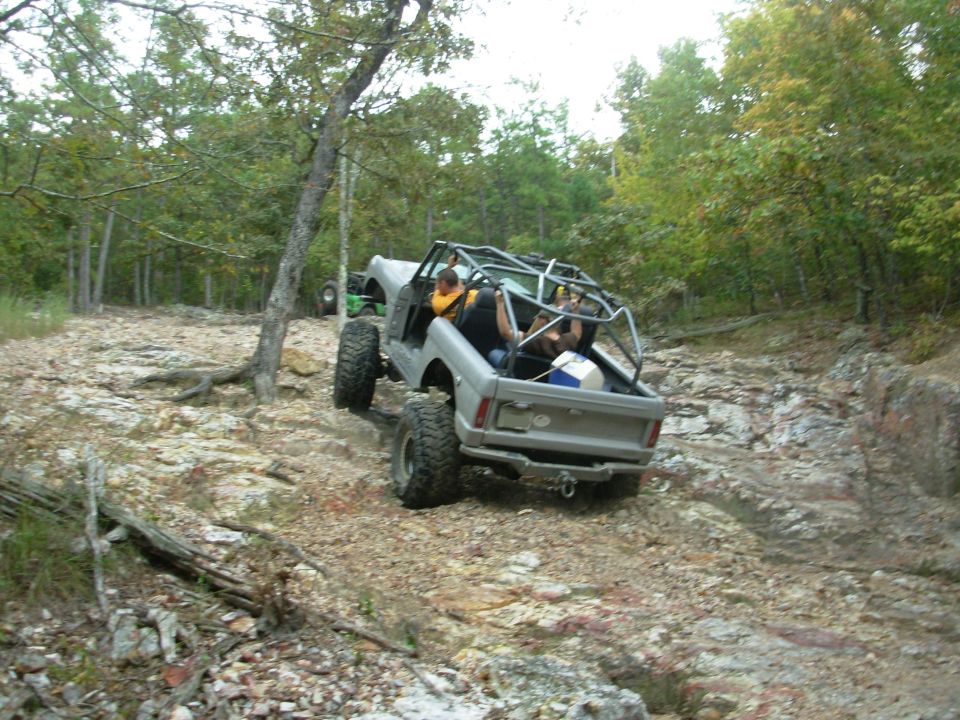 Silver open-top Bronco with roll cage descending a rocky wooded trail on mud tires.