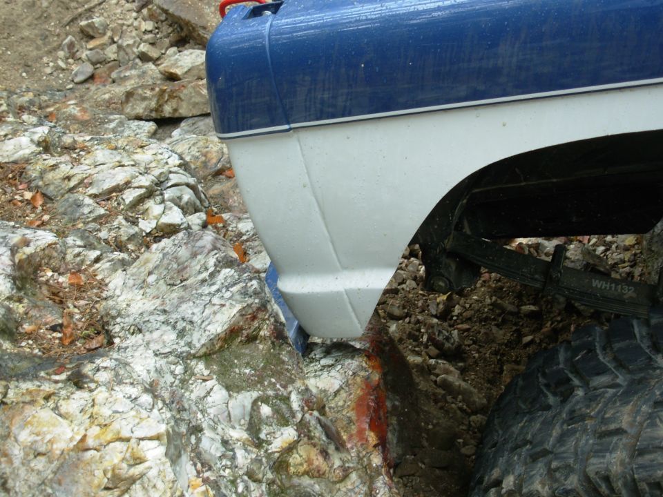 Blue and white truck fender pinched against a rock ledge, leaf spring and mud tire visible