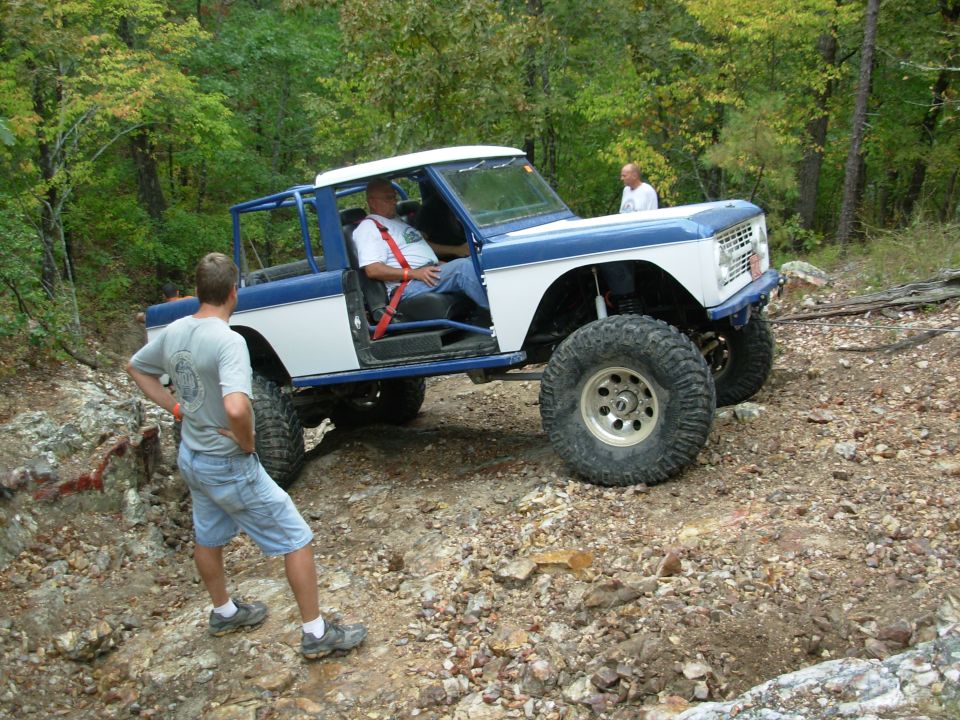 Blue and white early Ford Bronco with doors off and roll cage crawling rocky trail