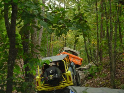 1983 Ford Bronco - Two off-road vehicles, including an orange 1983 Ford Bronco, navigate a challenging wooded trail.