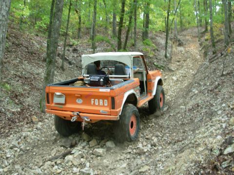 1983 Ford Bronco - Orange Ford Bronco navigates a challenging rocky incline on a wooded trail.