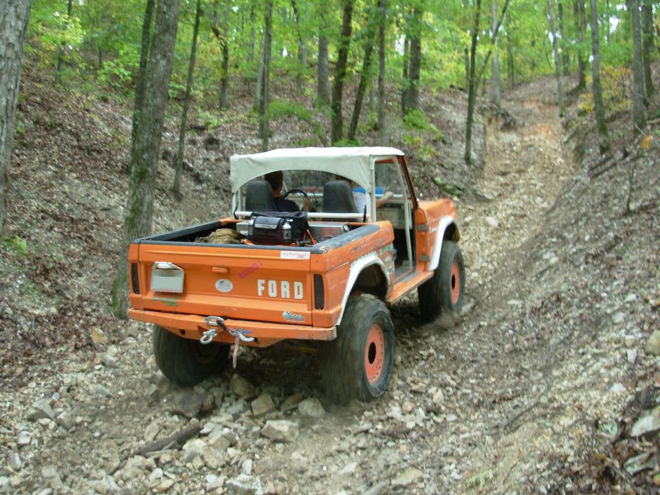Orange early Ford Bronco with white bikini top descending a rocky wooded trail, rear view.
