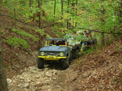 1983 Ford Bronco - Several off-road vehicles navigate a wooded trail with rocky terrain.
