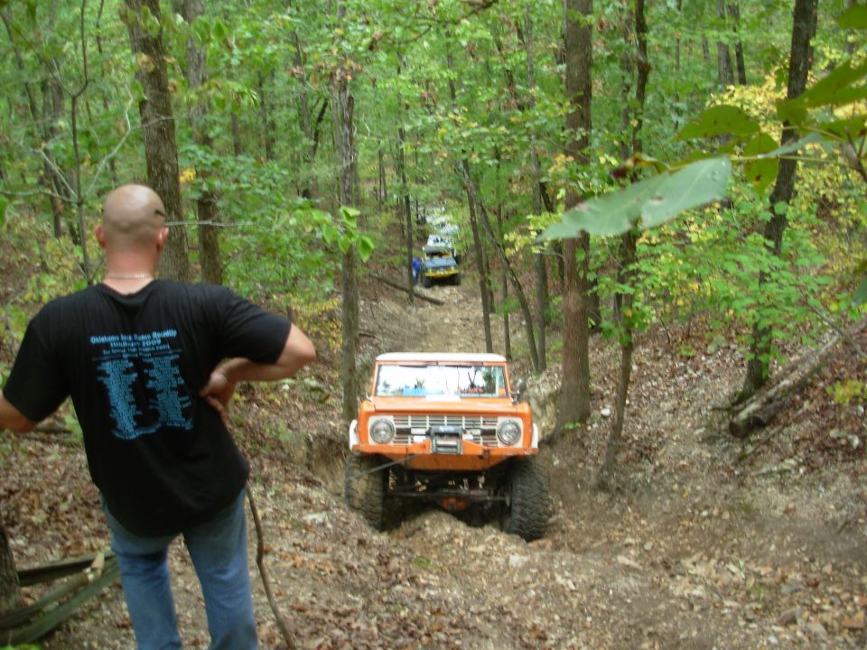 Orange lifted 4x4 with front winch climbing a rocky wooded trail as a spectator watches