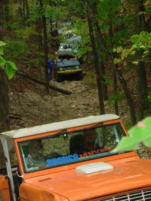 1983 Ford Bronco - An orange 1983 Ford Bronco, identified by its body style and a windshield sticker, leads a procession of off-road vehicles on a wooded trai…