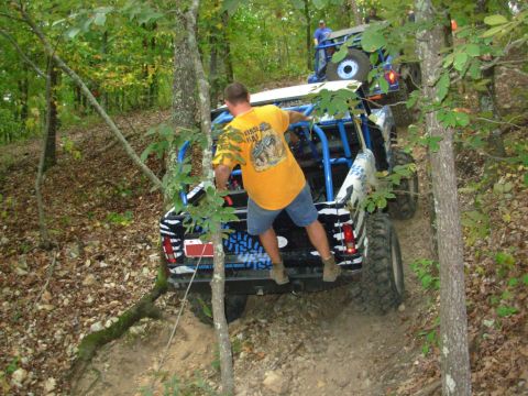 1983 Ford Bronco - Two off-road vehicles navigate a wooded trail with steep inclines and loose dirt.