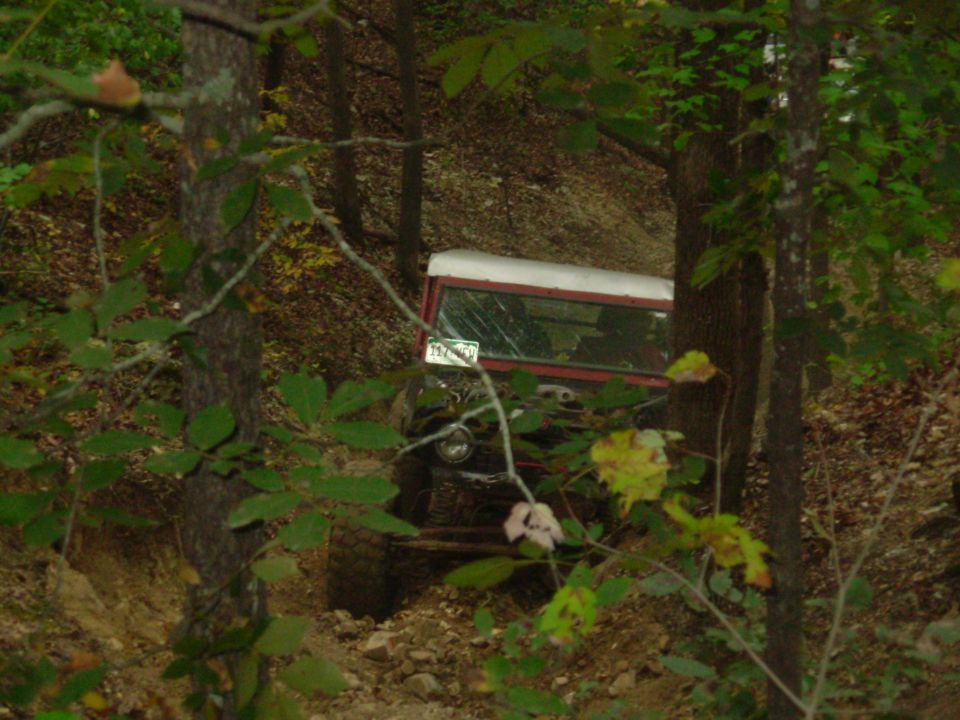 Red white-topped trail rig on steep rutted dirt bank seen through trees