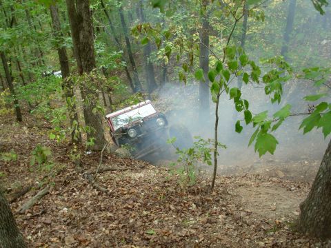 1983 Ford Bronco - A red off-road vehicle navigates a steep, wooded trail, kicking up dust and smoke.